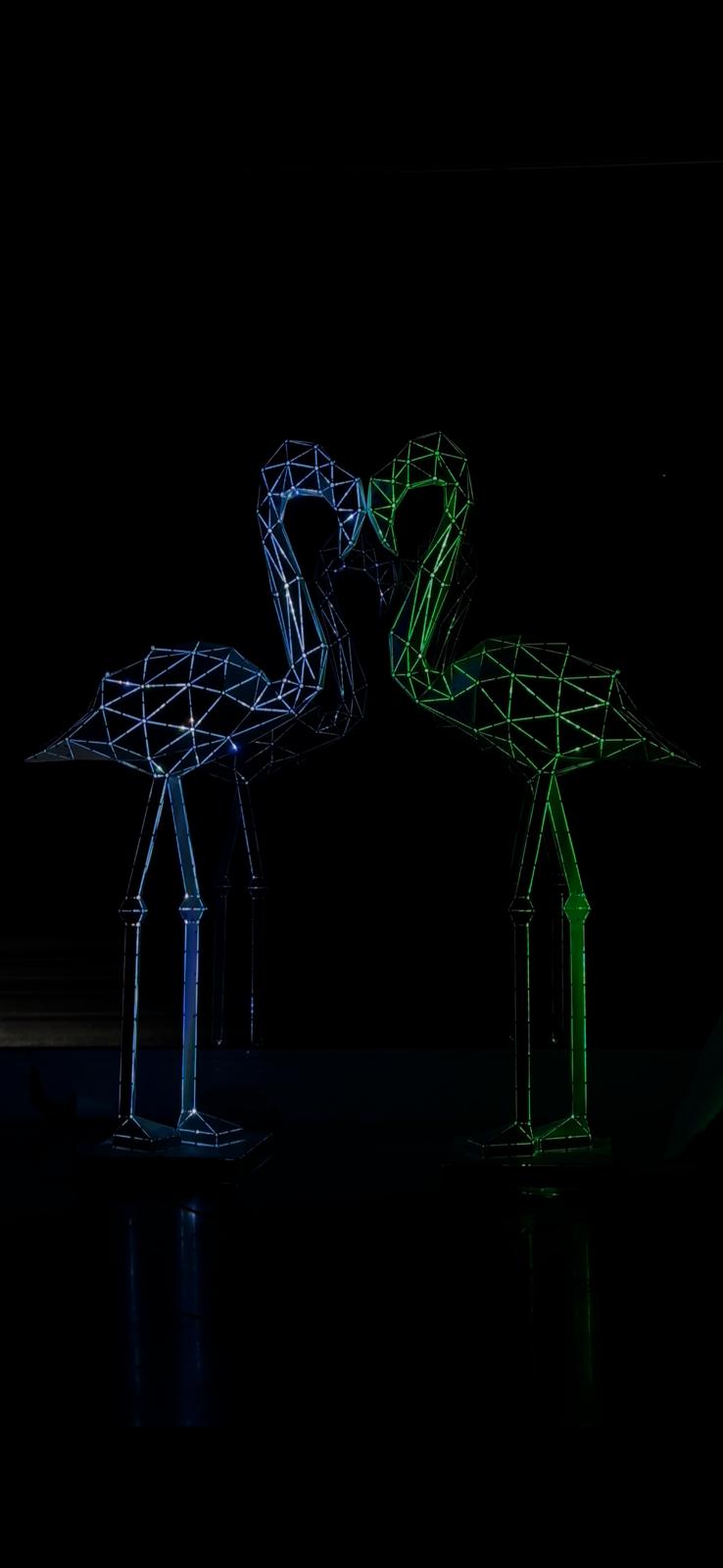 Two geometric metal flamingo sculptures in blue and green neon lighting against a black background