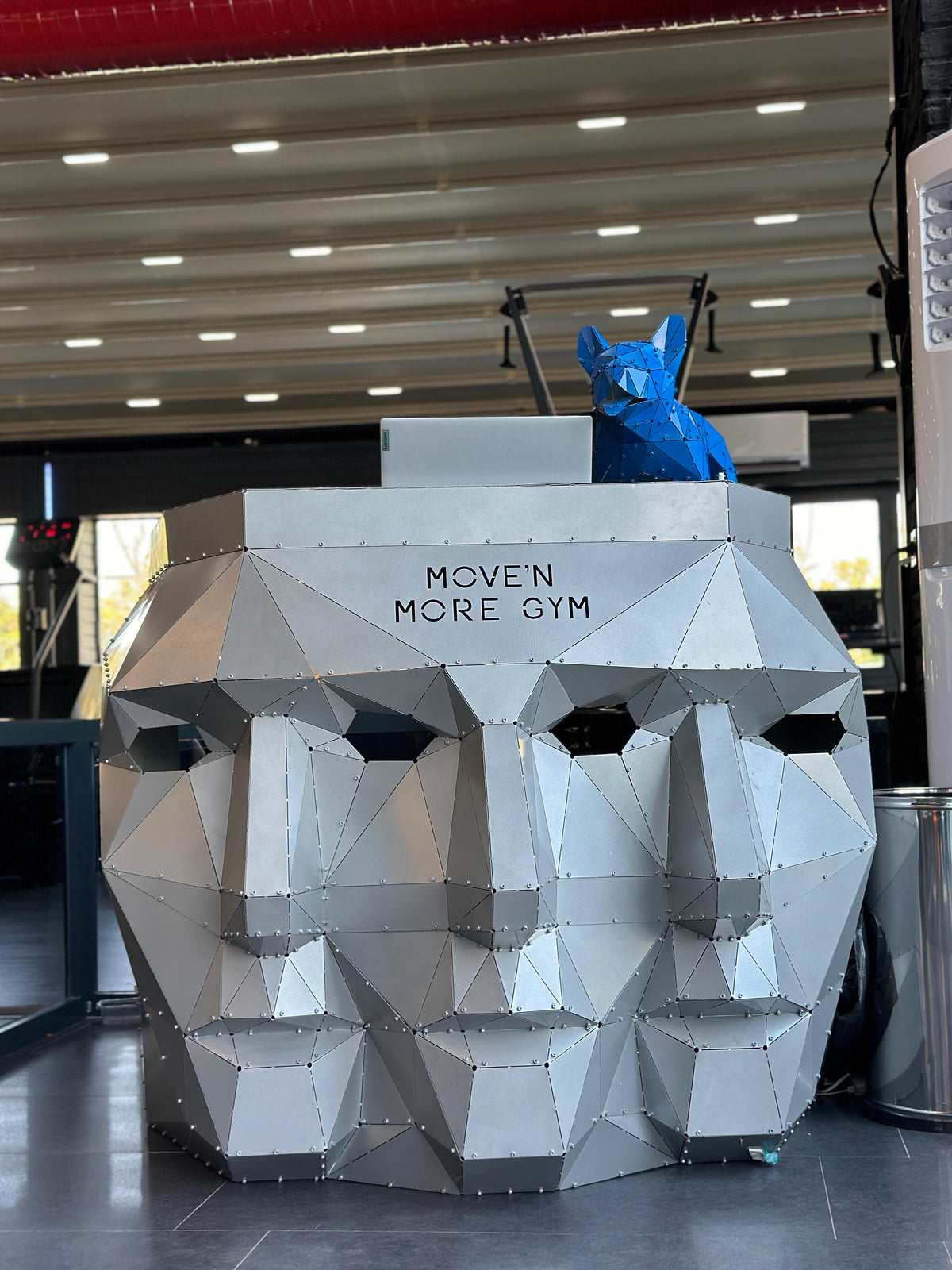 Low-poly silver metal head sculpture with MOVE'N MORE GYM text and a blue geometric fox on top.
