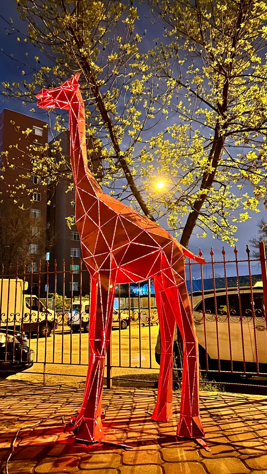 Red geometric metal giraffe sculpture illuminated at night in an urban setting