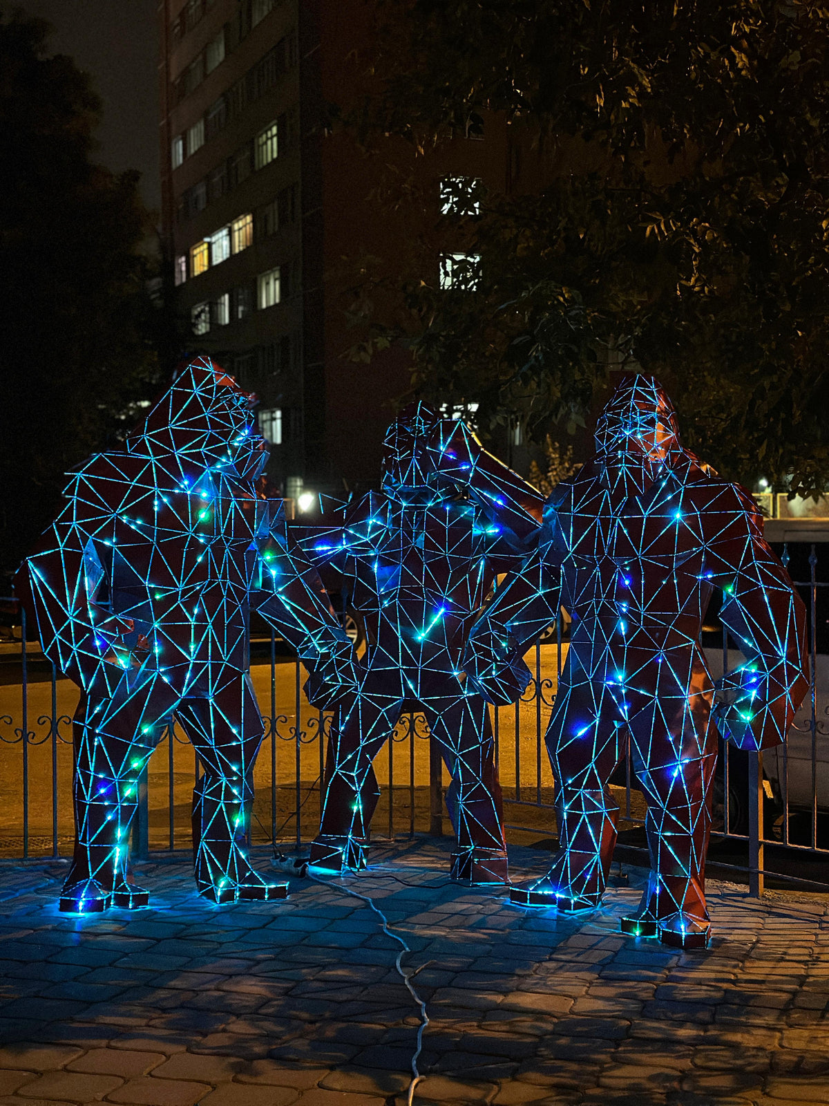 Three geometric metal gorilla sculptures illuminated with blue neon lights at night.
