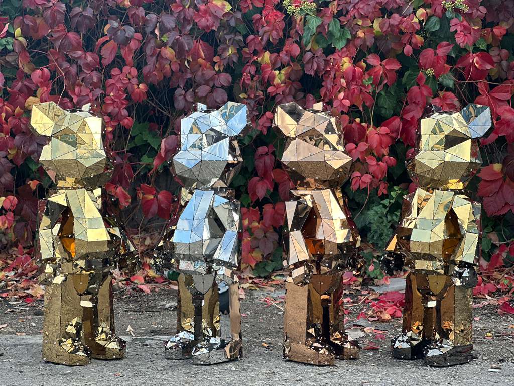 Four geometric faceted metal bear sculptures in gold and silver against autumn foliage.