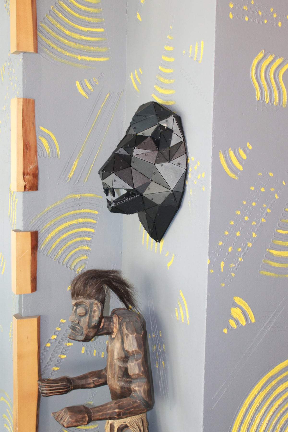 Geometric black metal lion head sculpture next to a wooden tribal mask on a textured grey wall