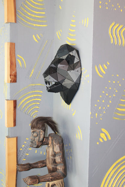 Geometric black metal lion head sculpture next to a wooden tribal mask on a textured grey wall