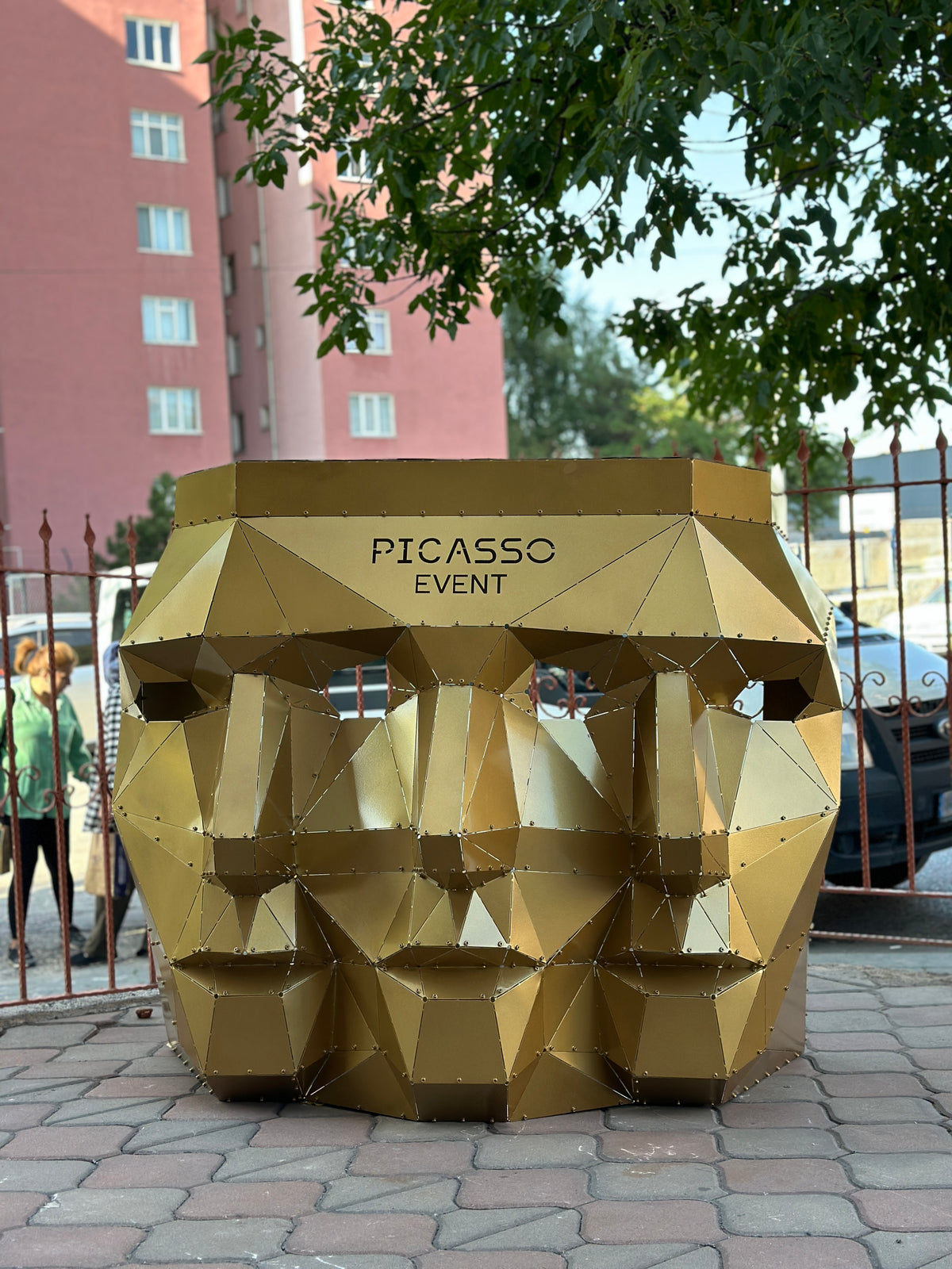 Geometric gold metal face sculpture with PICASSO EVENT text in an outdoor setting