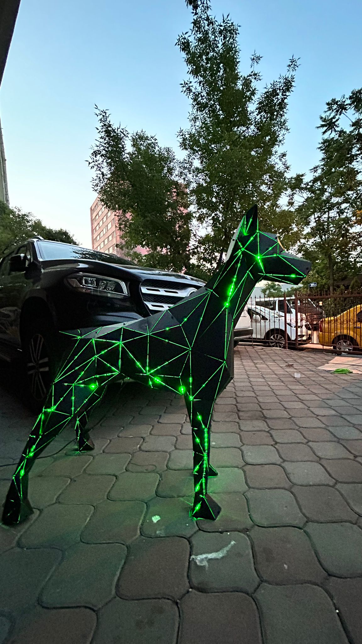 Geometric metal dog sculpture with glowing green LED lines on a paved outdoor area.
