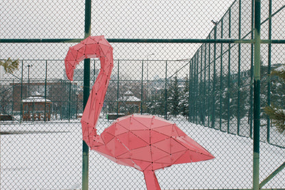 Geometric pink metal flamingo sculpture standing outdoors in a snowy field by a fence.