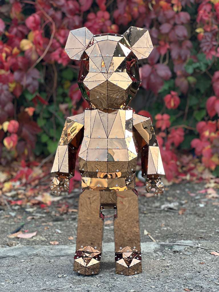Geometric polished copper bear sculpture standing outdoors against autumn foliage.