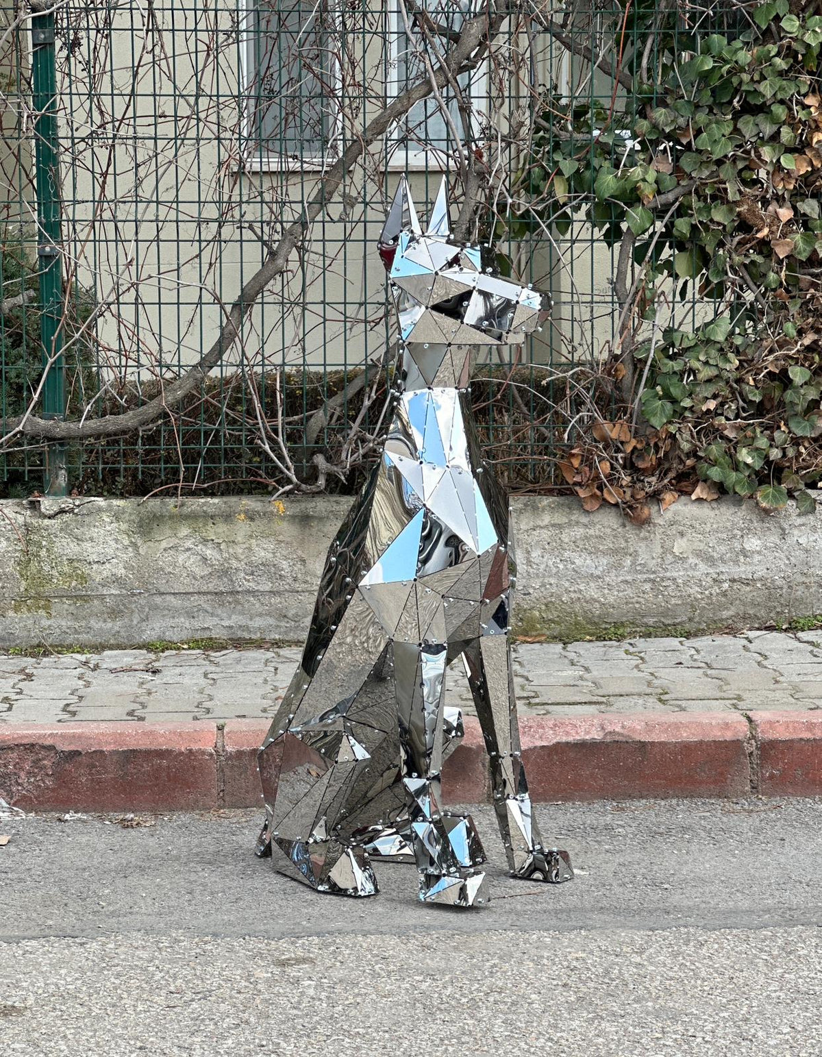 Geometric polished stainless steel dog sculpture sitting outdoors near a garden fence