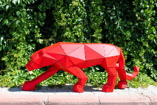 Geometric red metal panther sculpture standing against a lush green hedge background