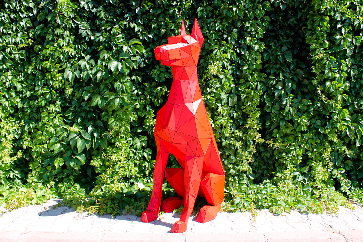Geometric red metal sculpture of a sitting dog against a lush green hedge background