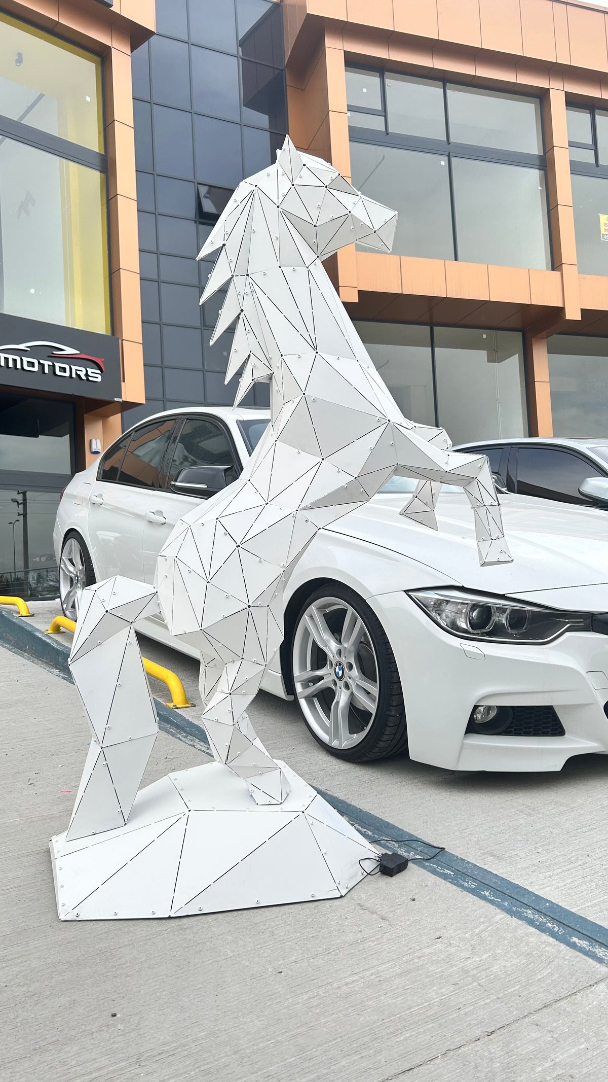 Geometric white metal horse sculpture standing next to a white luxury car.