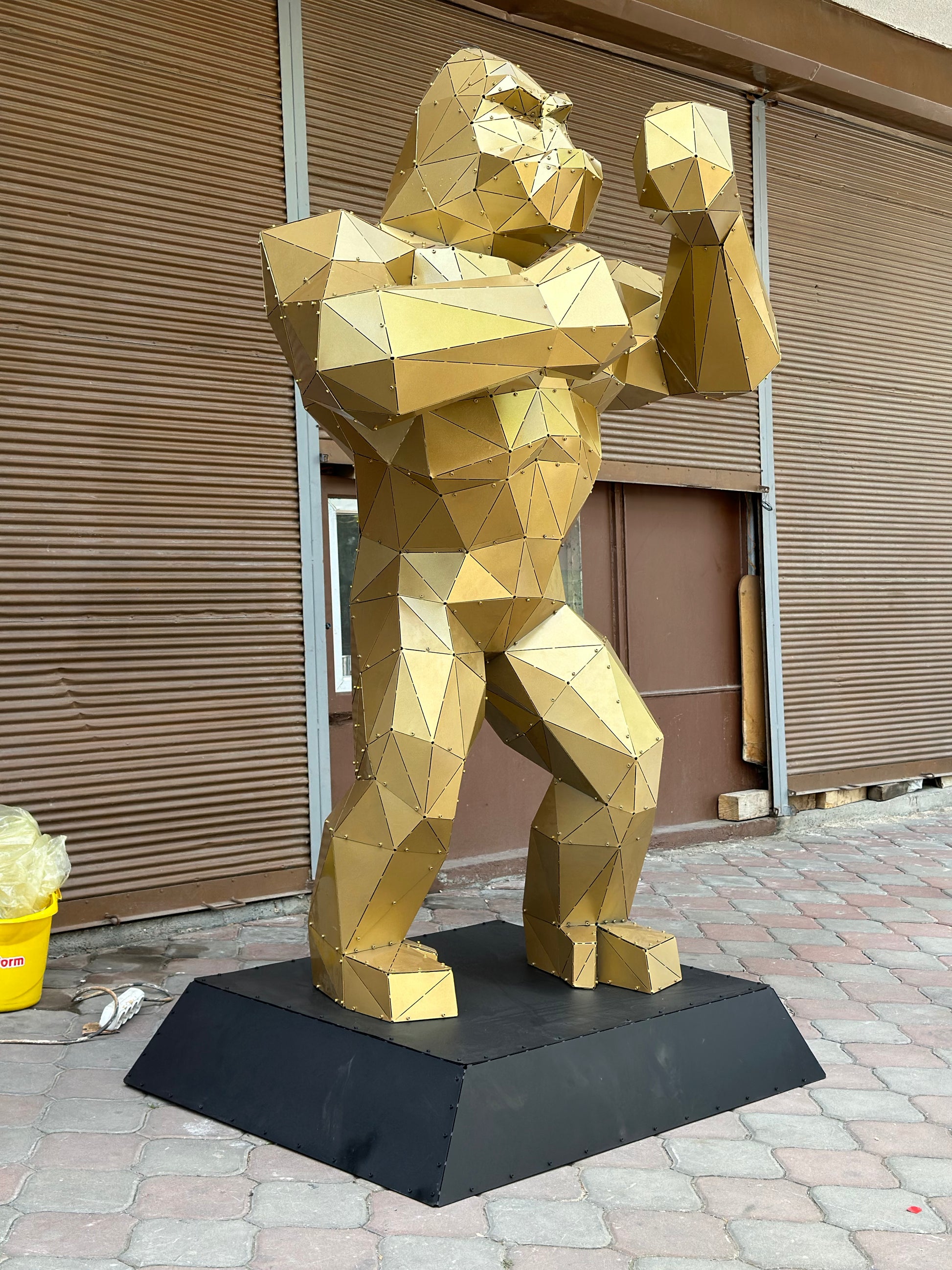 Gold low-poly metal gorilla sculpture standing on a black base outdoors with one fist raised
