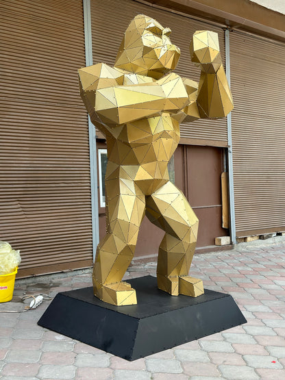 Gold low-poly metal gorilla sculpture standing on a black base outdoors with one fist raised
