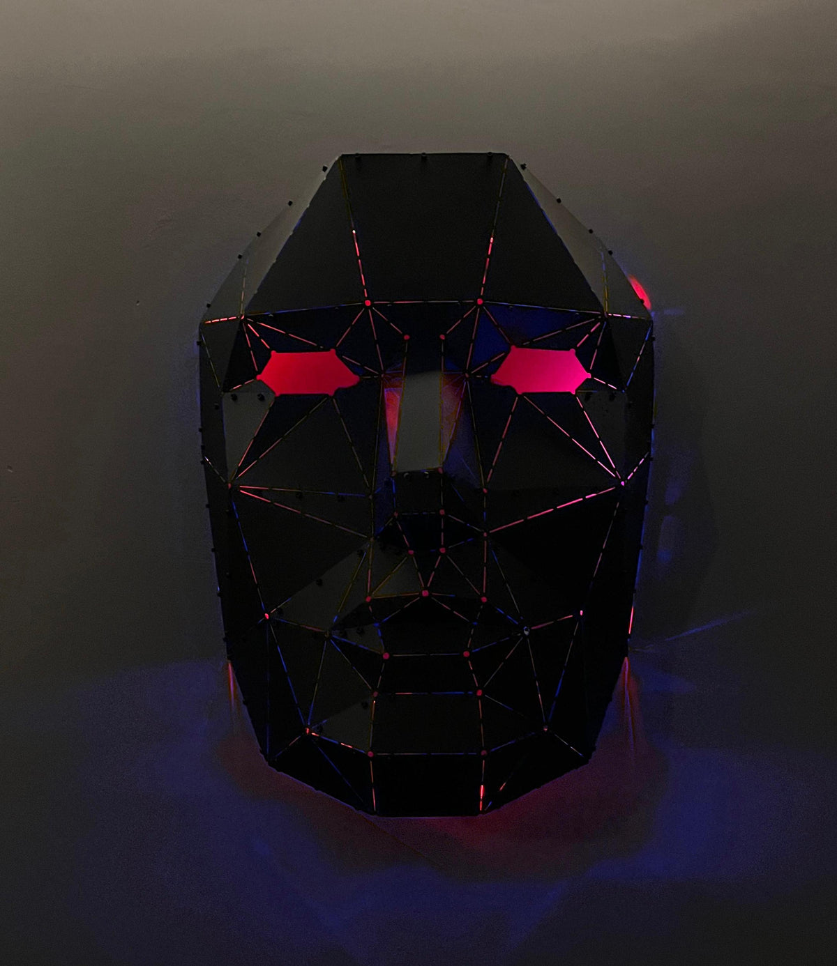 Handcrafted low-poly black metal face sculpture with glowing red eyes and pink accents.
