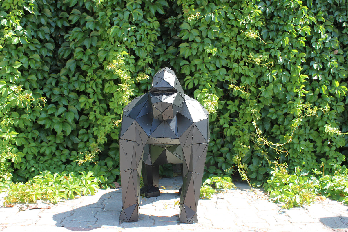 Handcrafted low-poly metal gorilla sculpture positioned against a lush green hedge background
