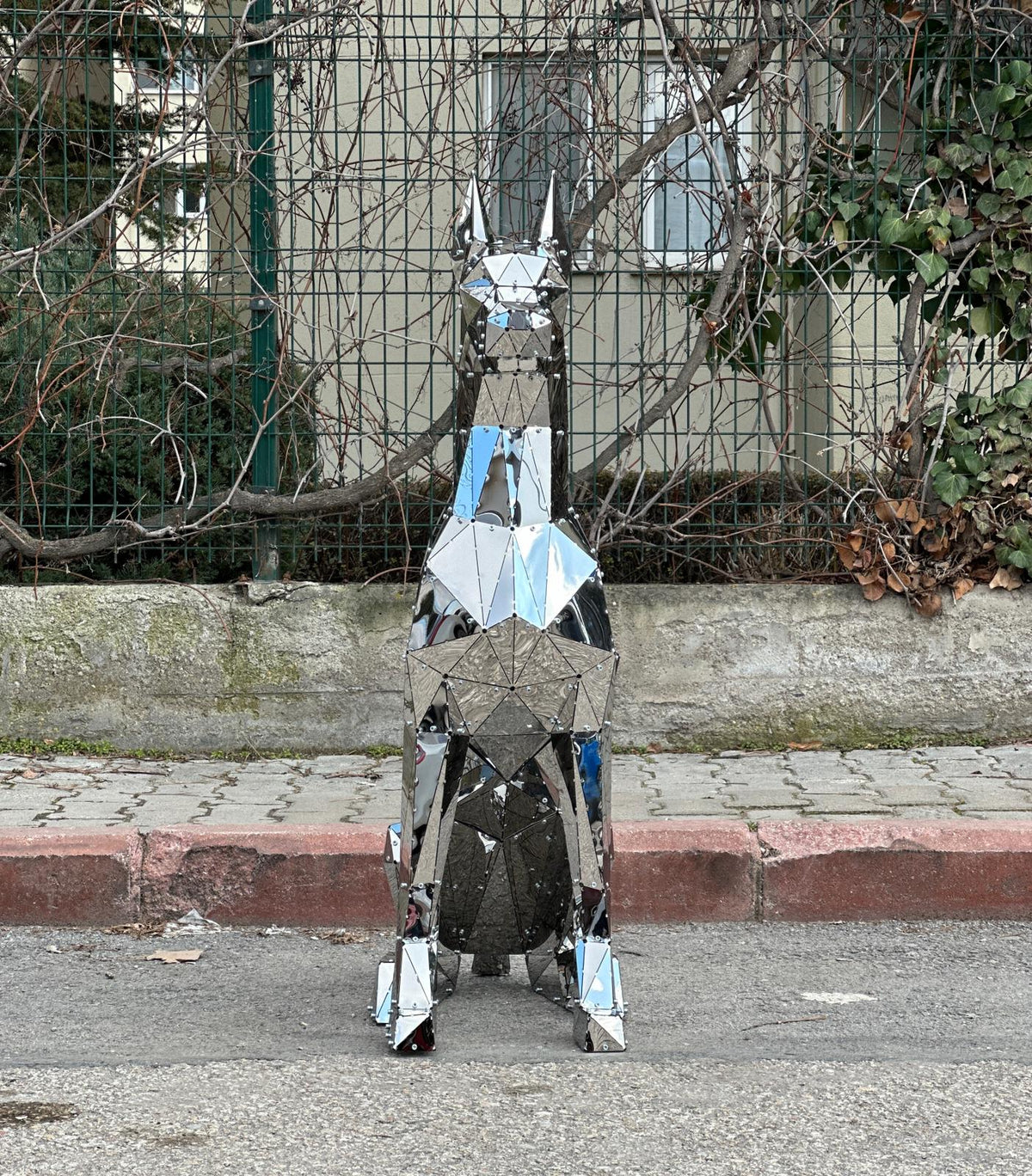 Handcrafted mirrored metal dog sculpture with geometric facets on a city sidewalk