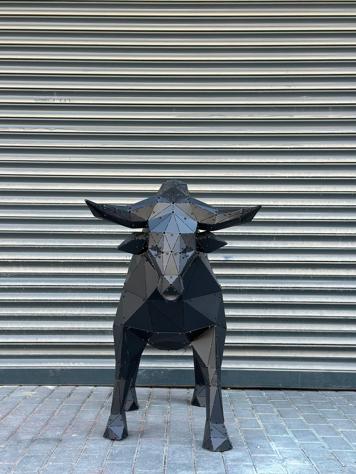 Low-poly black metal bull sculpture standing against a gray ribbed industrial shutter.