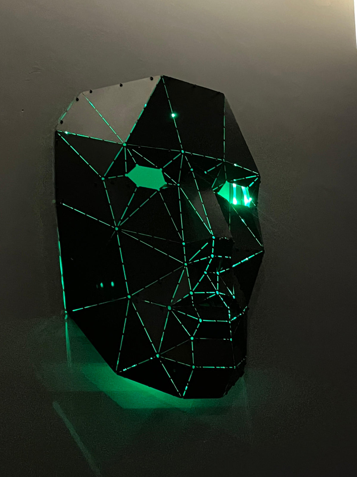 Low-poly black metal human face sculpture with glowing green neon light accents.
