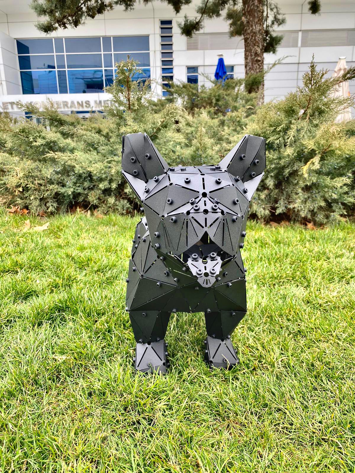 Low-poly black metal French Bulldog sculpture standing on a green grass lawn.