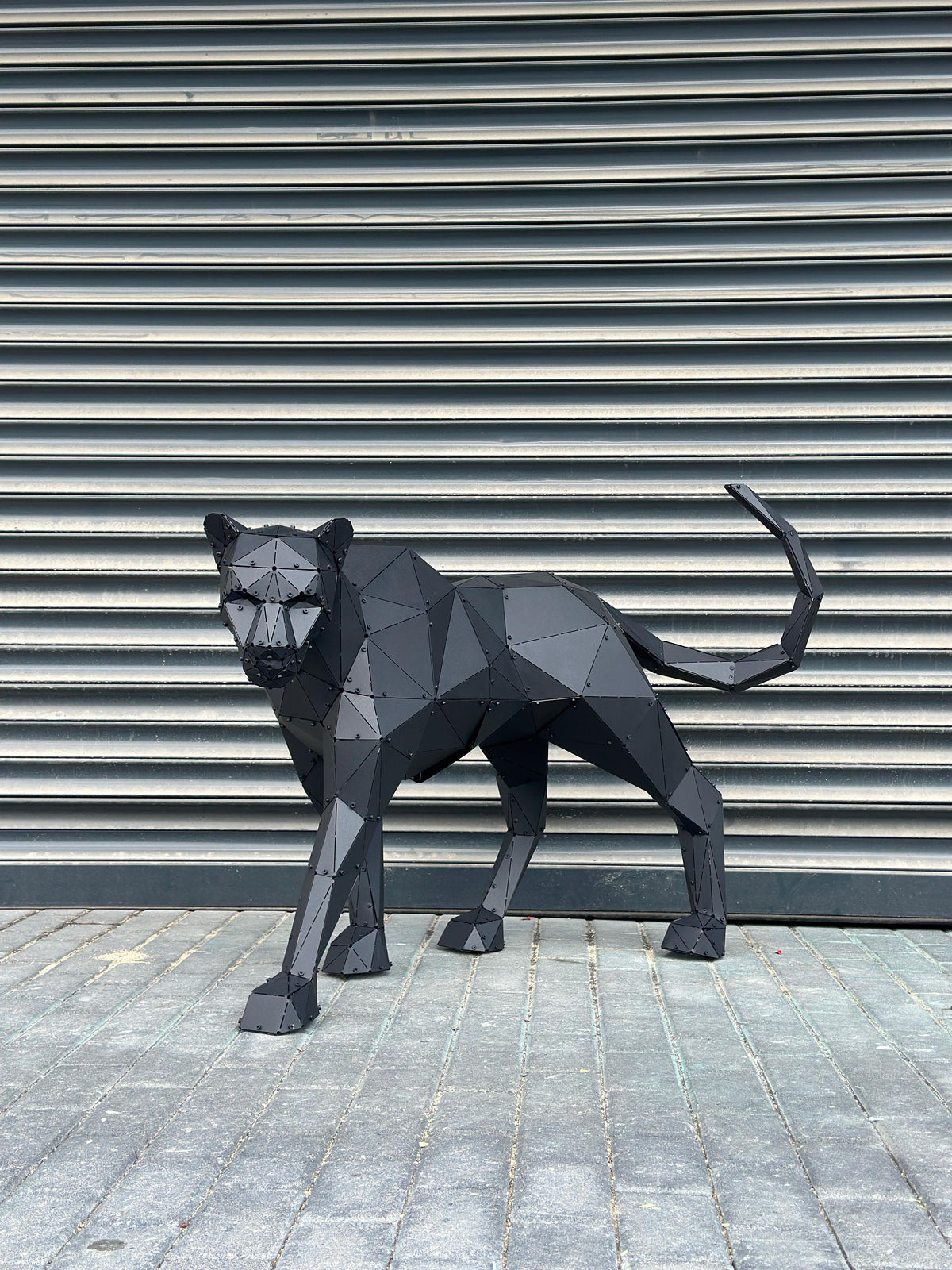 Low-poly black metal panther sculpture standing against a grey industrial shutter background