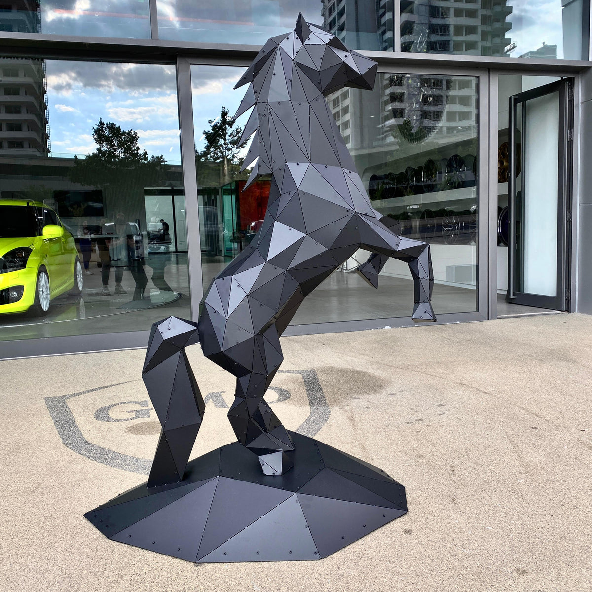 Low-poly black metal horse sculpture rearing on a concrete surface in front of a modern building.