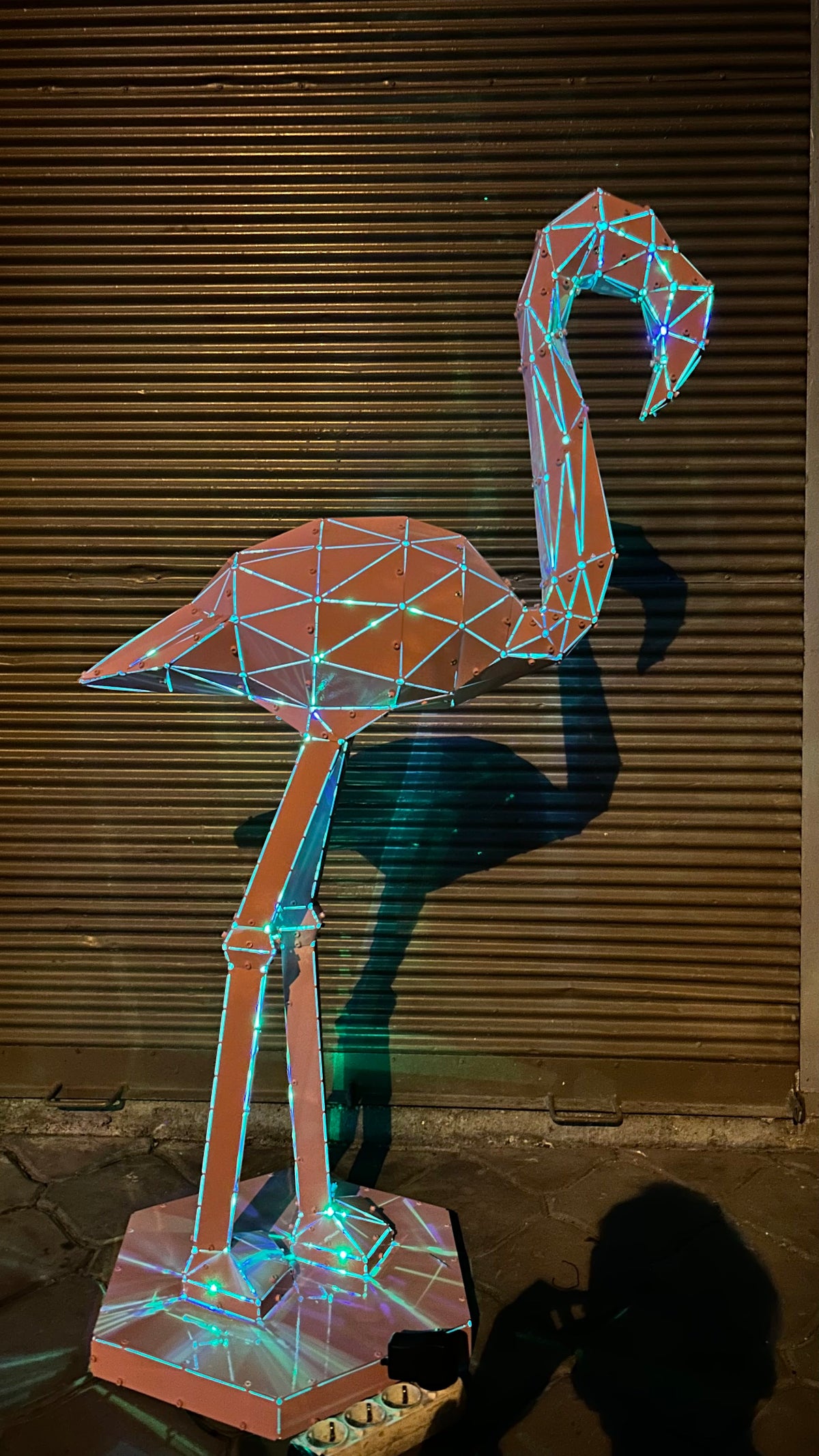 Geometric low-poly flamingo metal sculpture with integrated neon lighting against a shutter background
