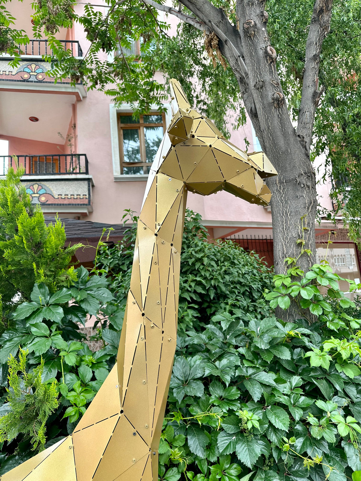 Low-poly gold metal giraffe sculpture standing in a lush garden near a pink building.
