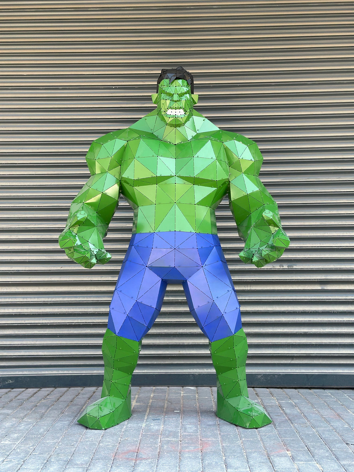 Low-poly metal sculpture of the Hulk in green and blue against a grey shutter background.