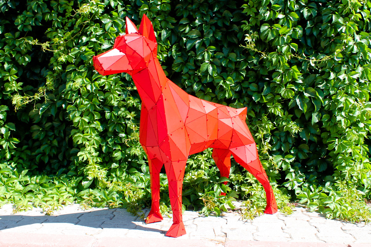 Low poly red metal dog sculpture standing against a lush green hedge.
