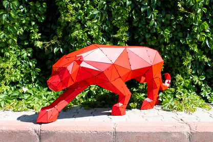 Low-poly red metal panther sculpture standing against a lush green hedge background