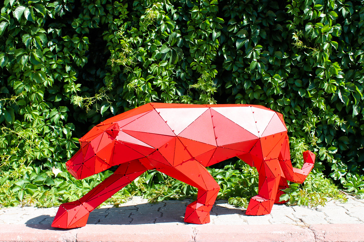 Low-poly red metal panther sculpture standing against a lush green hedge background.