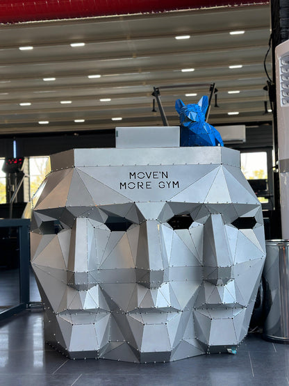 Low-poly silver metal head sculpture with MOVE'N MORE GYM text and a blue geometric fox on top.