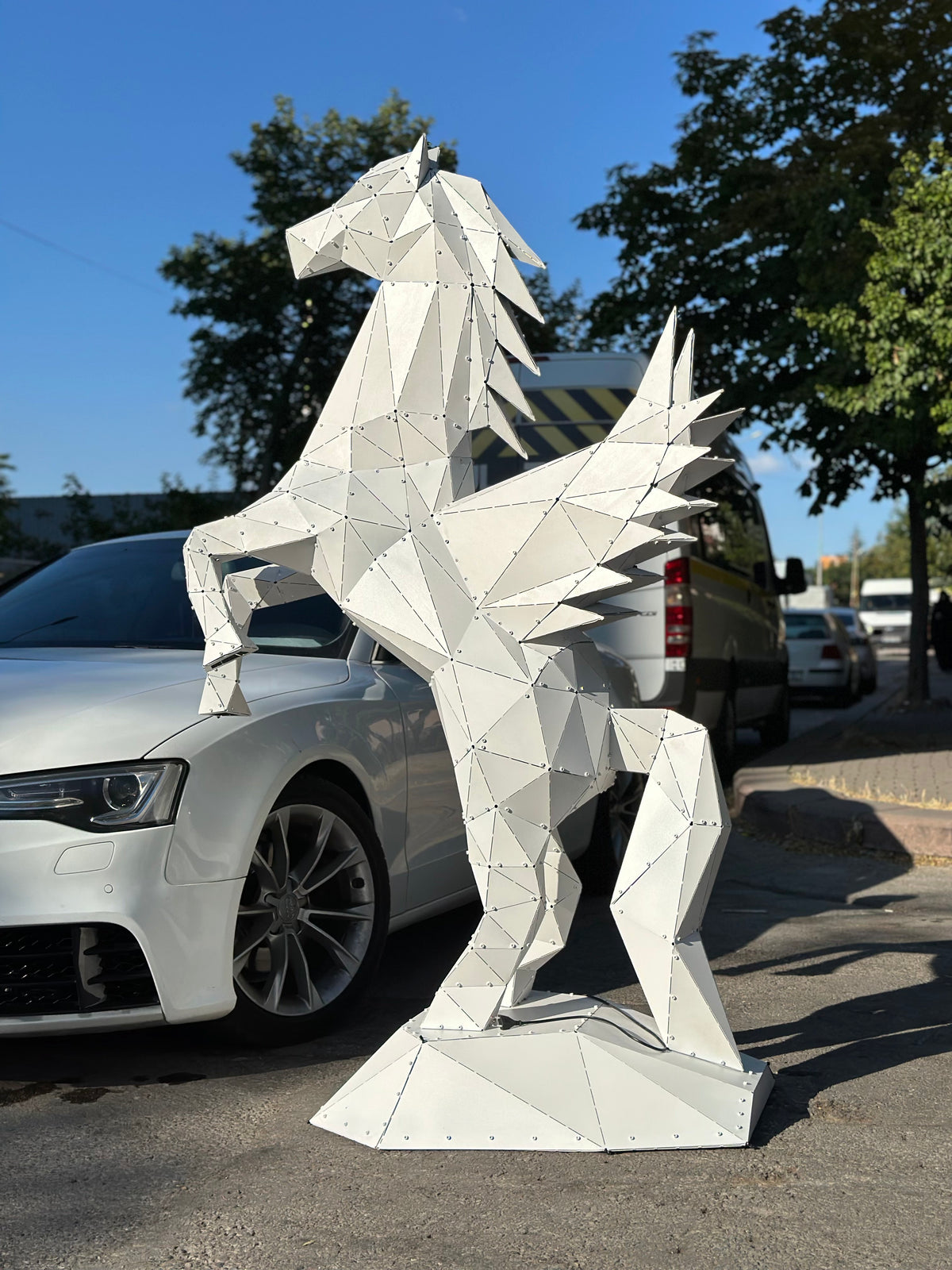 Low-poly white metal Pegasus sculpture standing next to a white car outdoors.