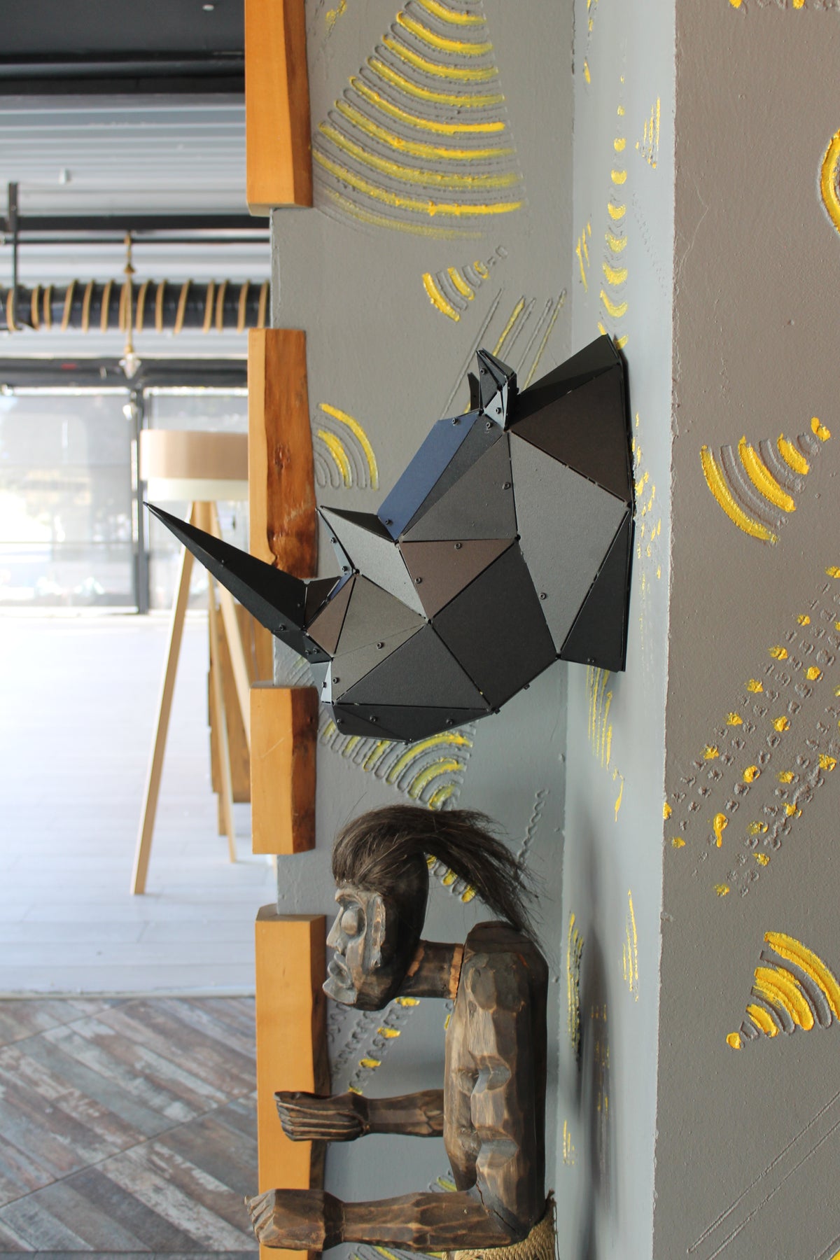 Modern geometric black metal wall sculpture and carved wooden figure on a grey textured wall.