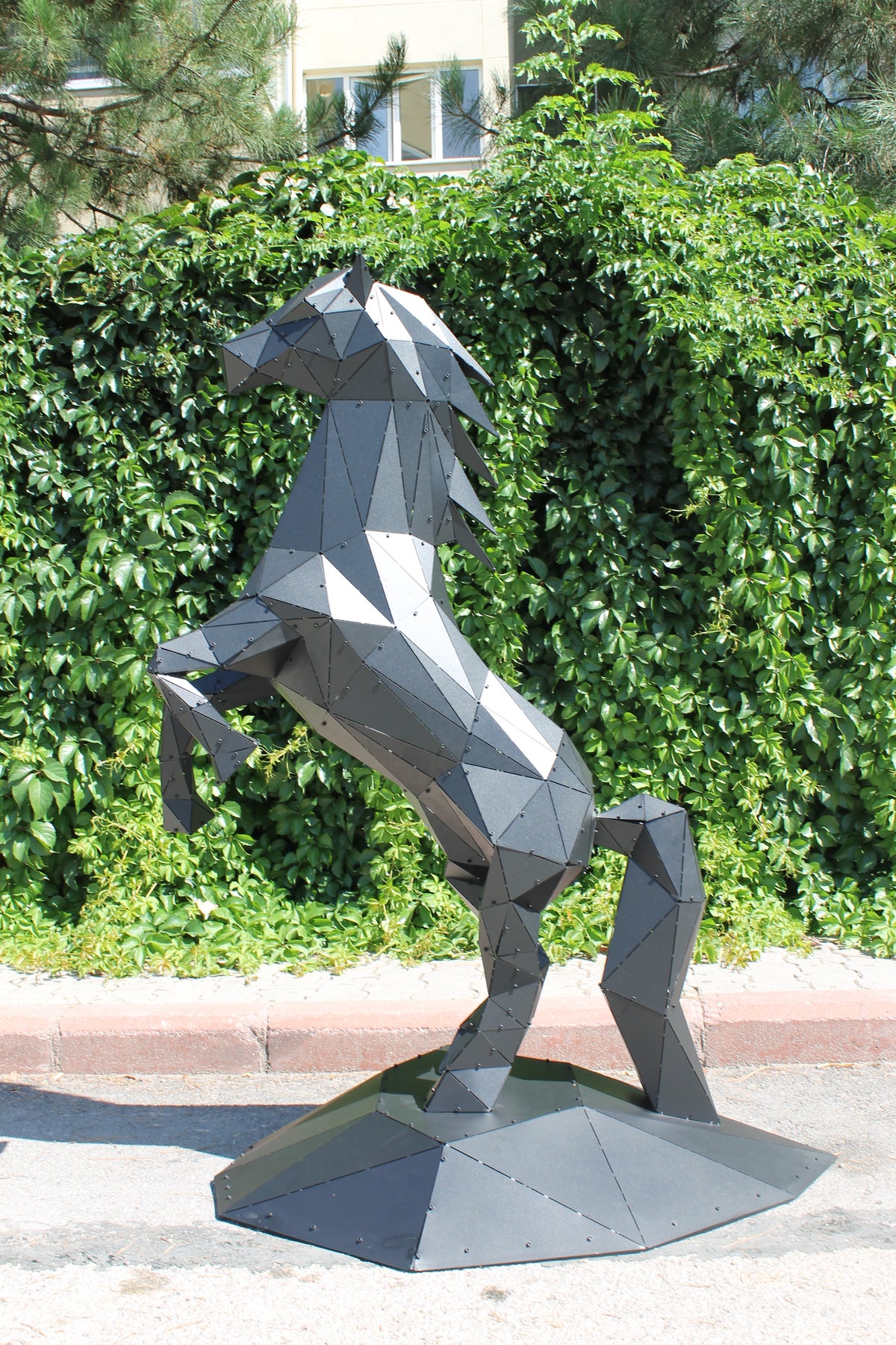 Modern low-poly metal horse sculpture rearing up against a lush green hedge background.