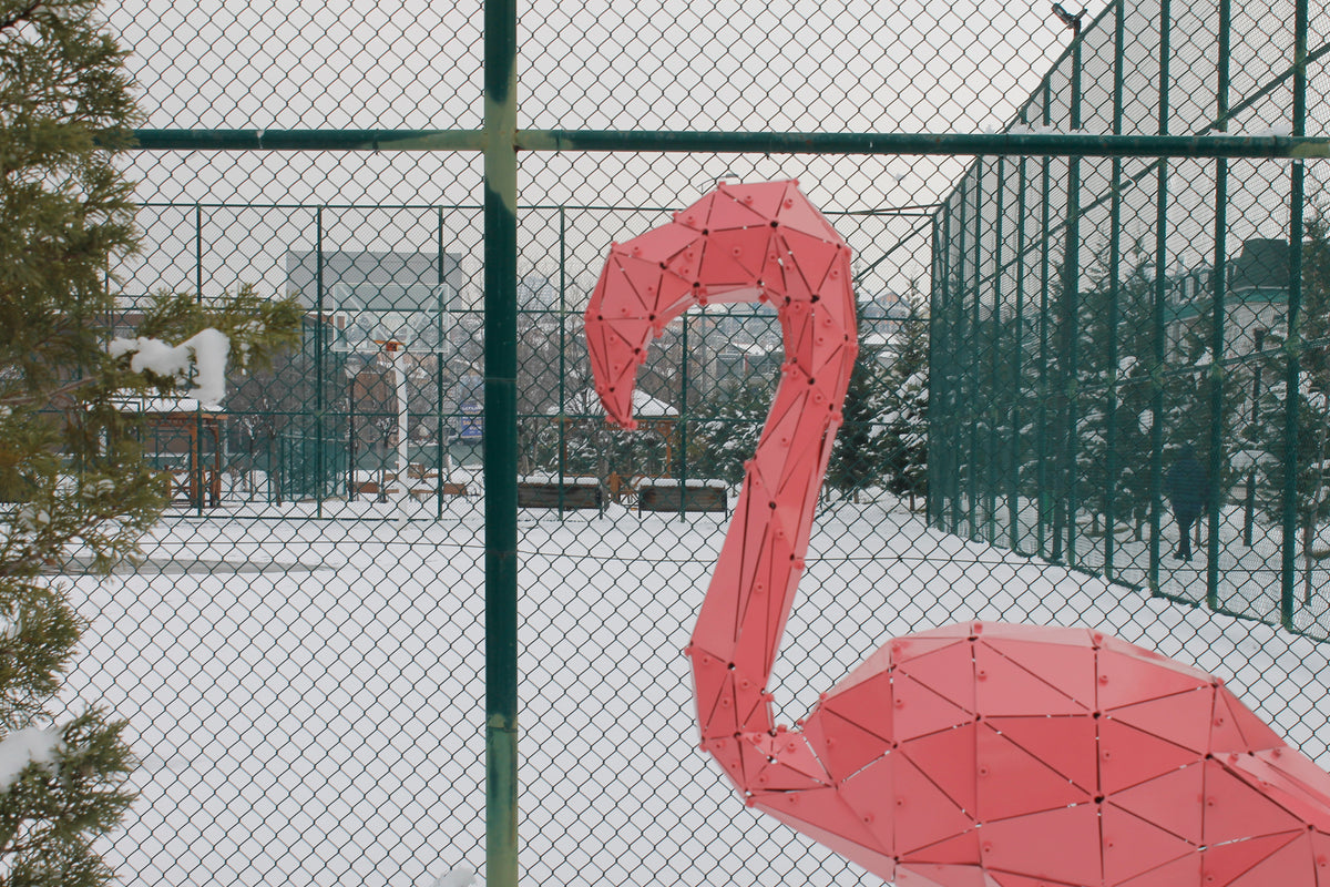 Pink geometric metal flamingo sculpture in a snowy outdoor setting with a chain-link fence.