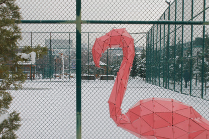 Pink geometric metal flamingo sculpture in a snowy outdoor setting with a chain-link fence.