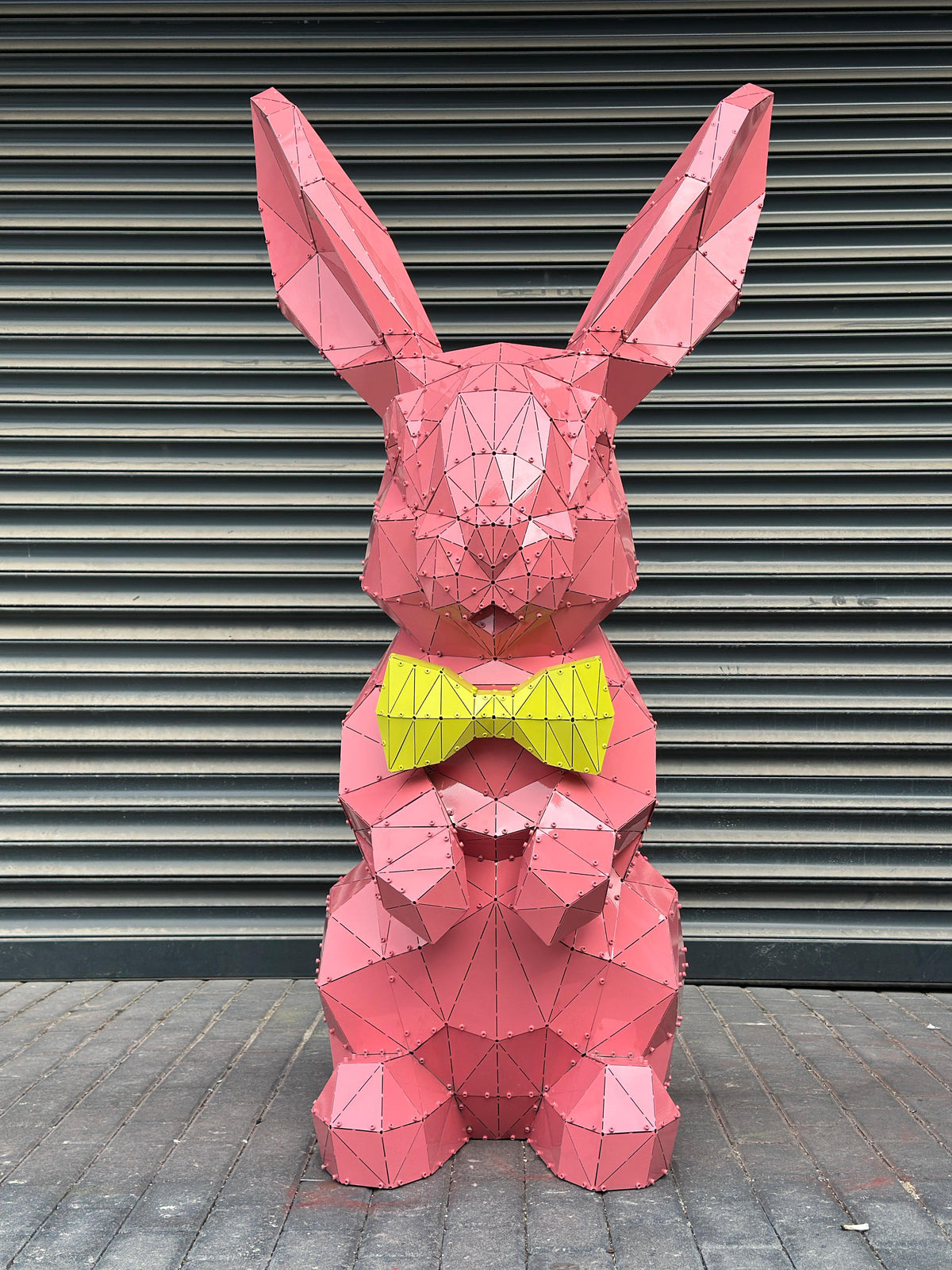 Pink low-poly metal rabbit sculpture wearing a yellow bow tie against a grey shutter.