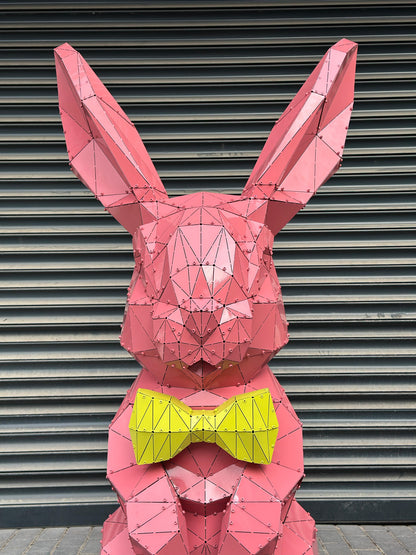Pink geometric metal rabbit sculpture wearing a yellow bowtie against a grey shutter background