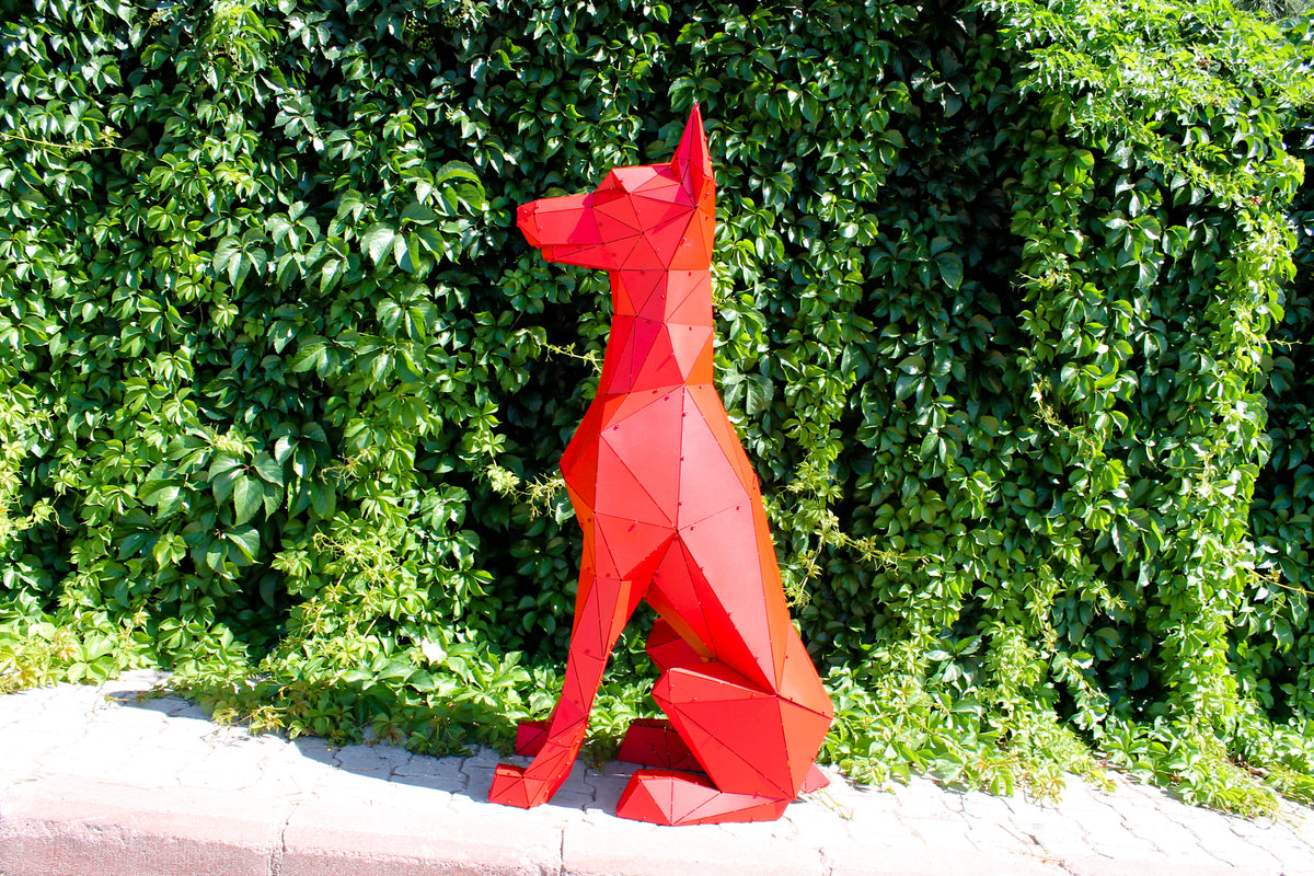 Red geometric low-poly Doberman sculpture sitting against a lush green hedge, side view
