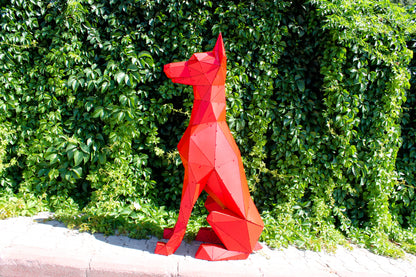 Red geometric low-poly Doberman sculpture sitting against a lush green hedge, side view