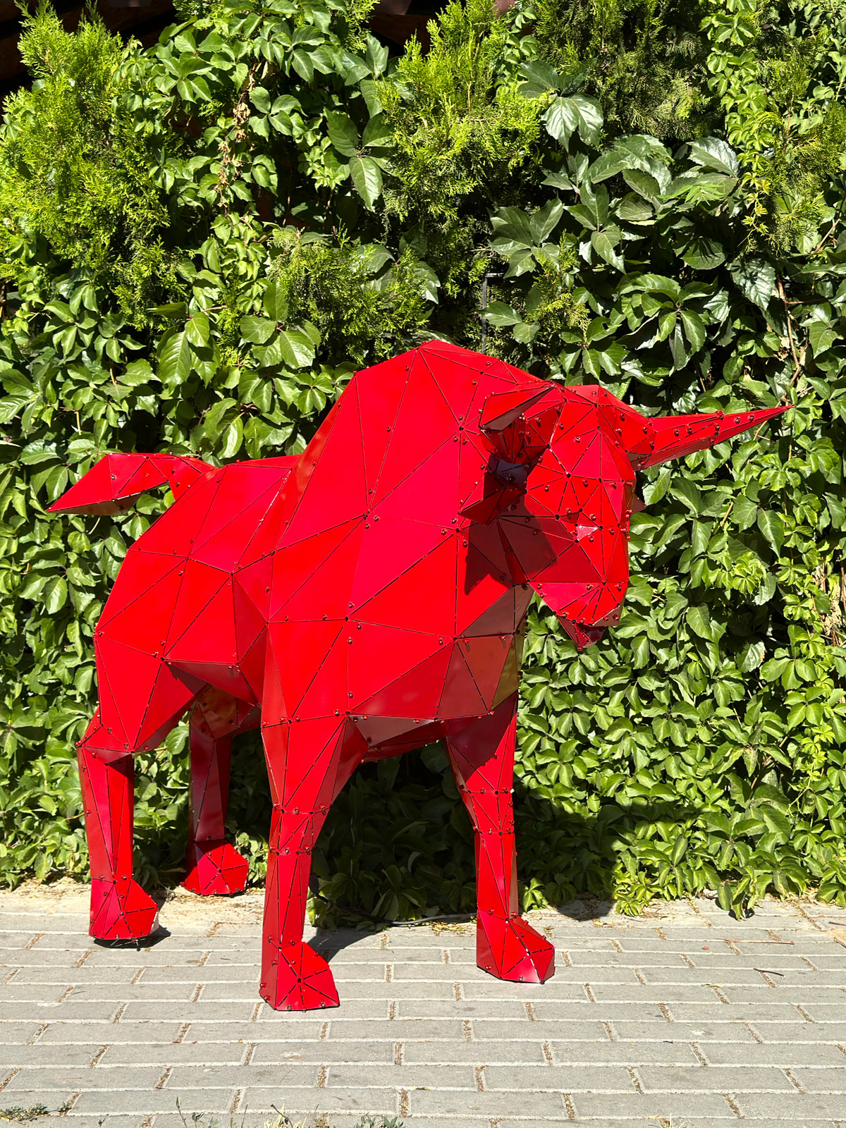Red geometric metal bull sculpture standing against a lush green hedge backdrop