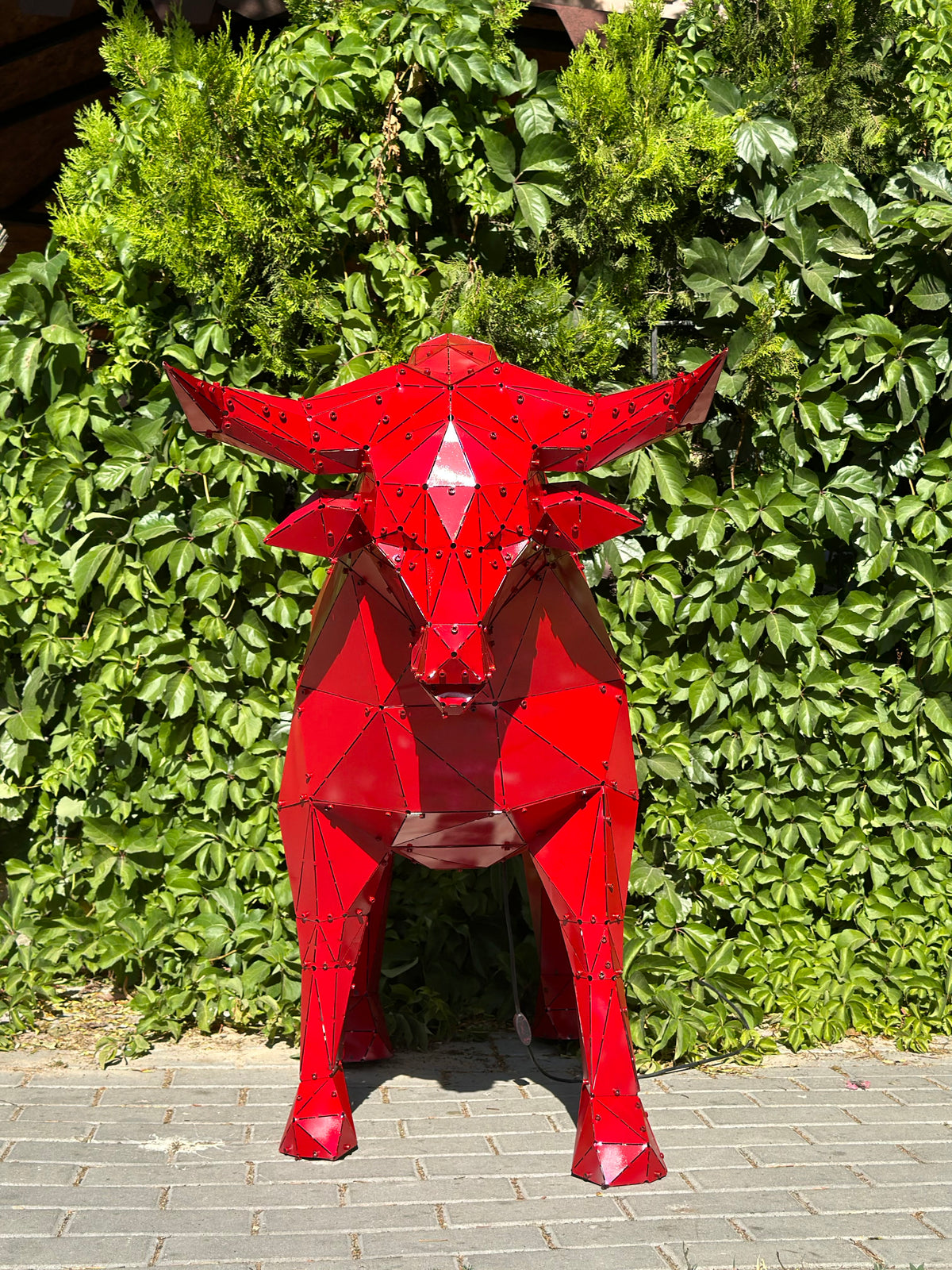 Red geometric metal bull sculpture standing against a lush green hedge background