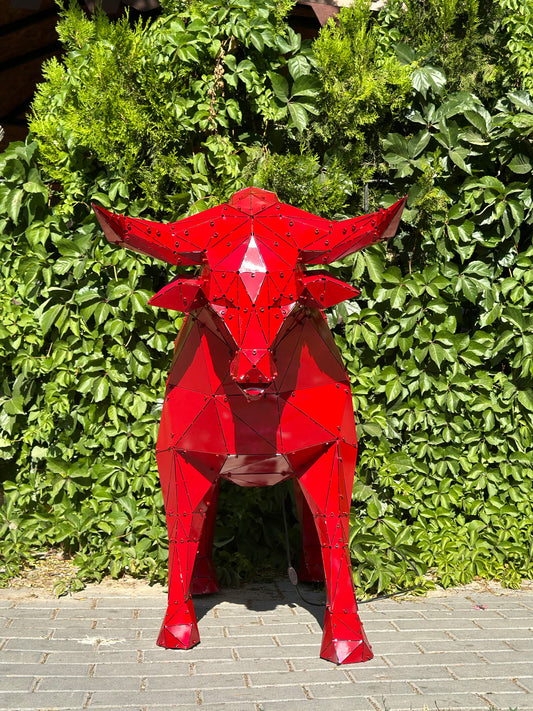 Red geometric metal bull sculpture standing against a lush green hedge background