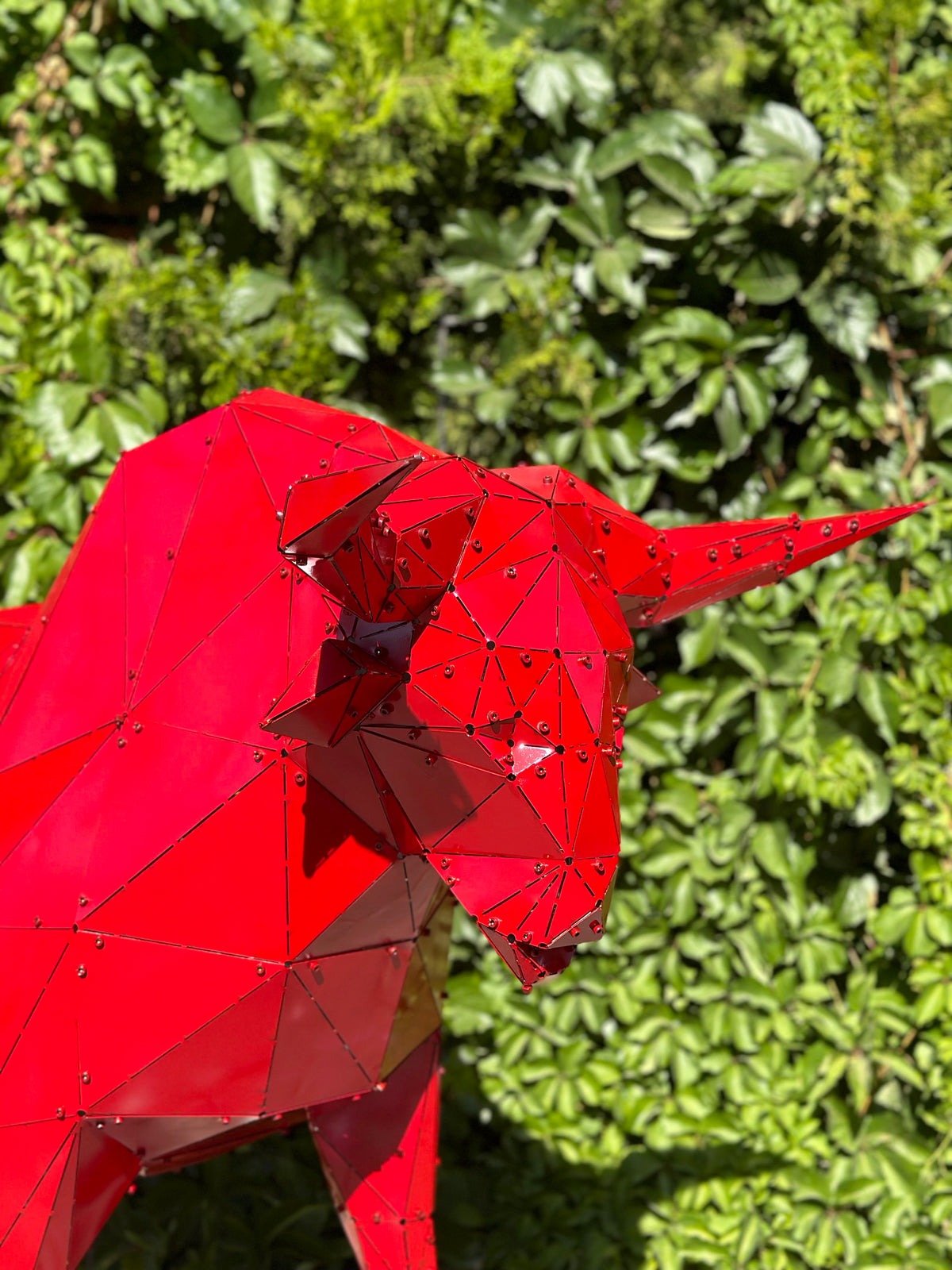 Red geometric metal bull sculpture against a lush green hedge background