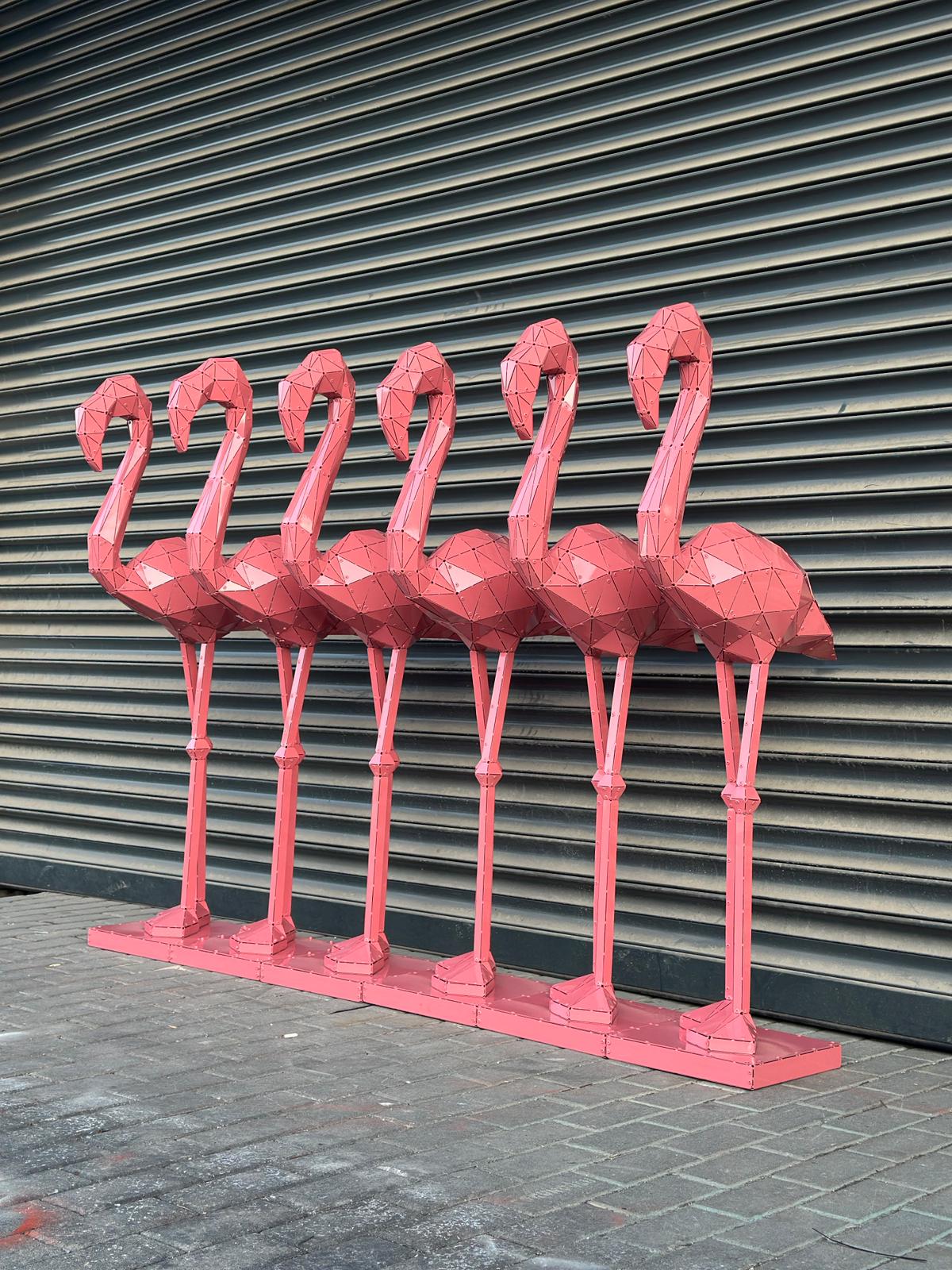 Six pink geometric metal flamingo sculptures standing against a grey shutter background.