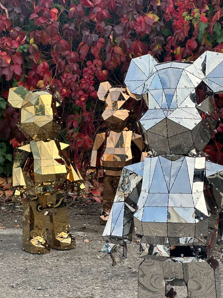 Three geometric metal bear sculptures in gold, bronze, and silver finishes against red foliage.