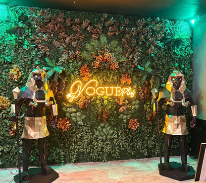 Two geometric metal sculptures of pharaohs flanking a neon Vogueplus sign against a greenery wall.