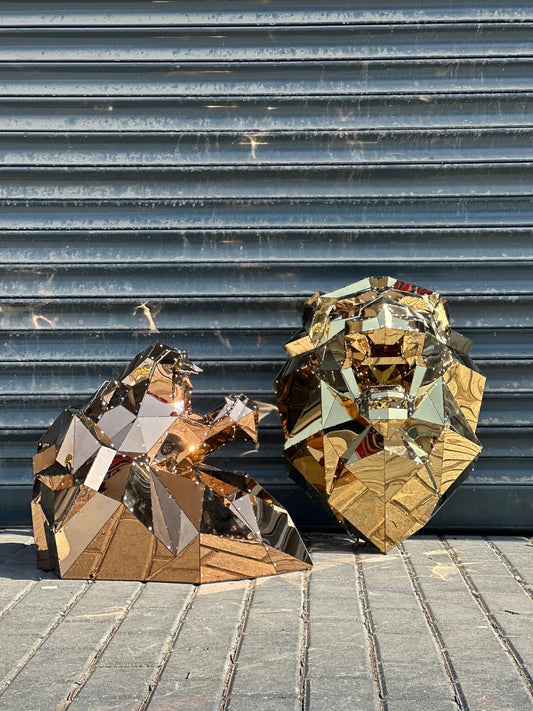 Gold mirrored geometric lion head and gorilla head sculptures on the ground against a grey metal shutter