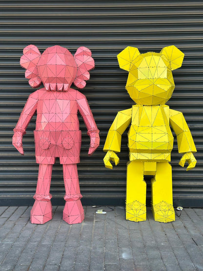 Two large, low-poly metal bear sculptures in pink and yellow against a grey shutter wall.